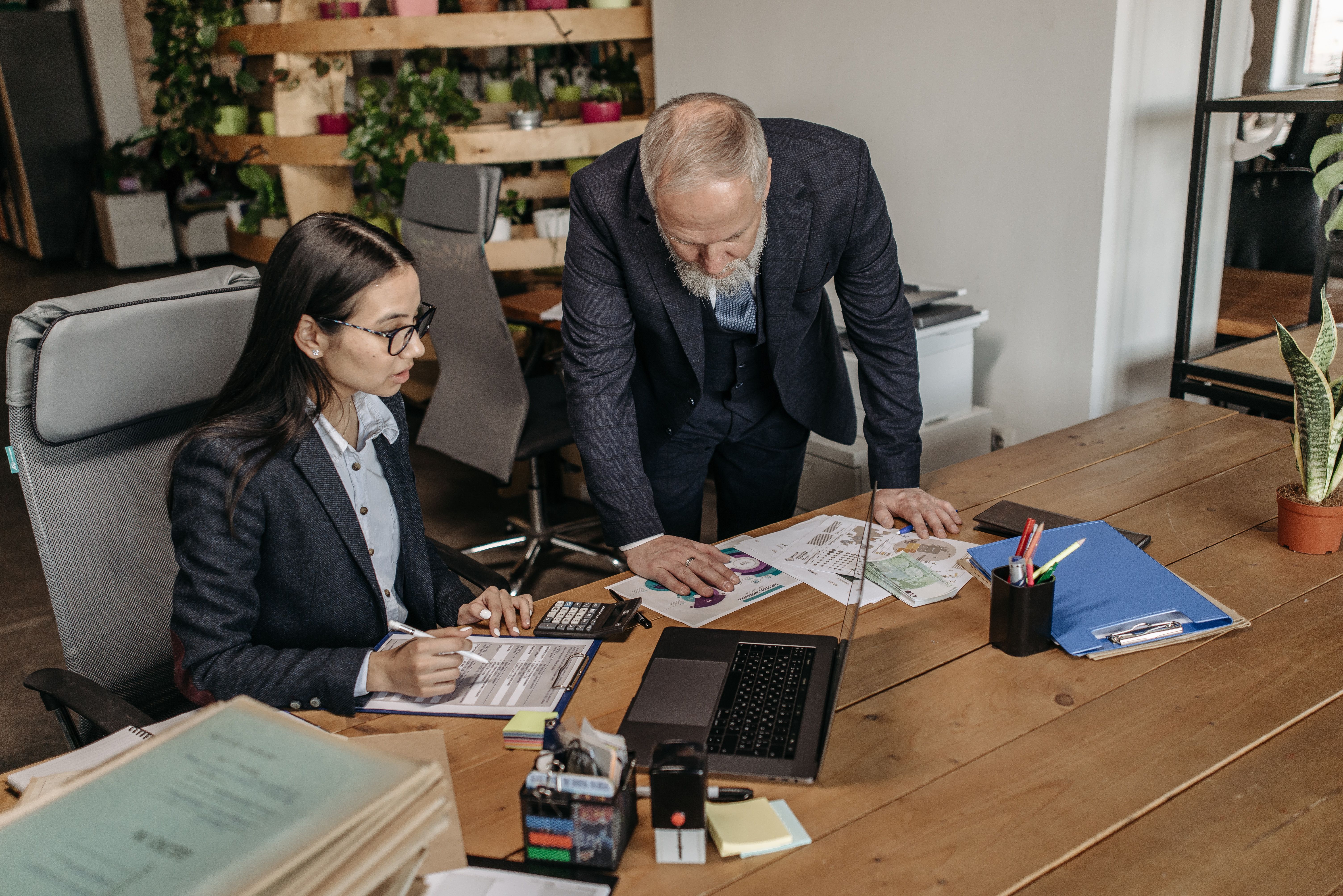A seated woman fills out a form while a standing man leans on a desk covered with charts and papers. | Source: Pexels