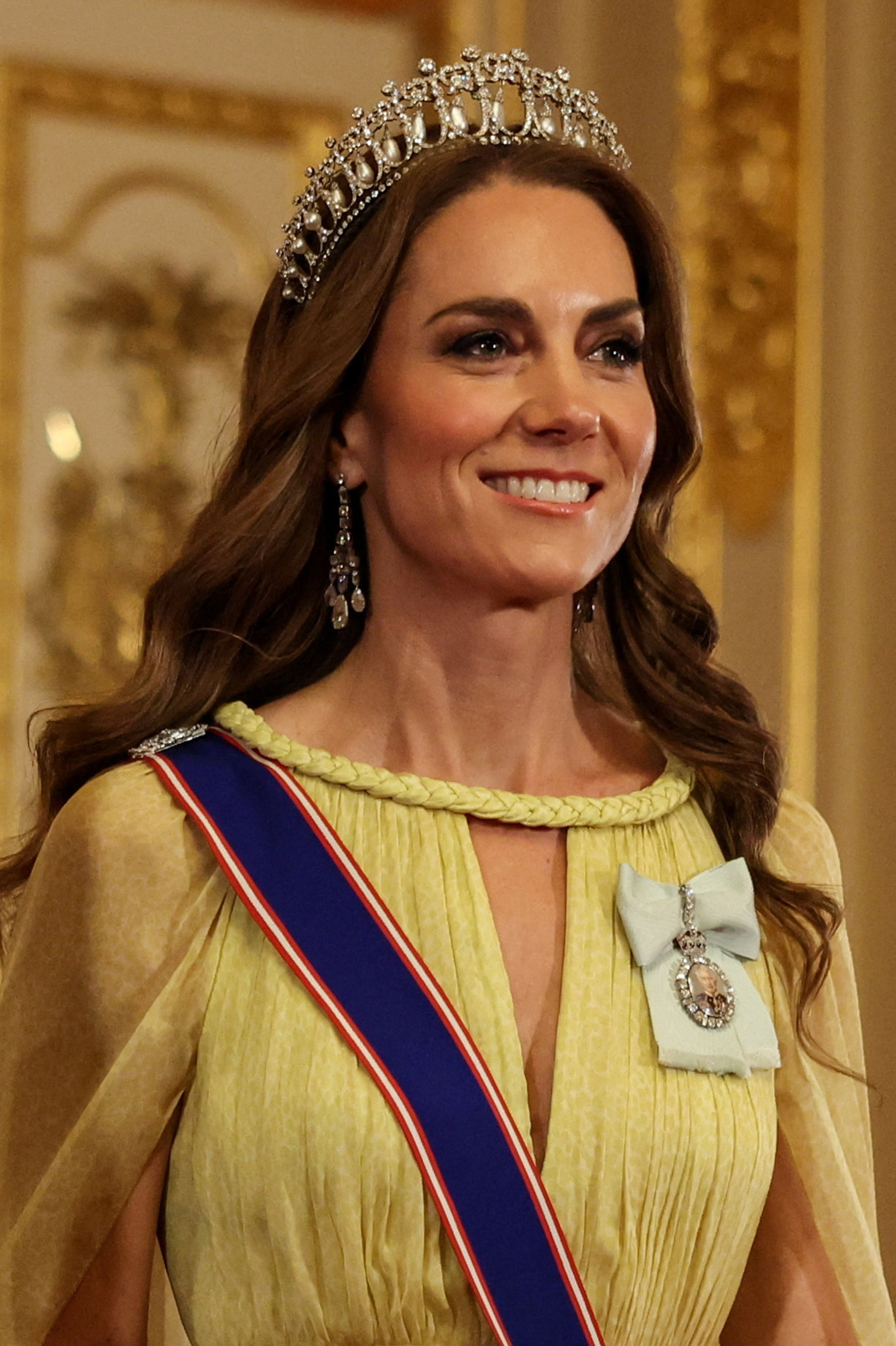 A detailed close-up captures Catherine, Princess of Wales, displaying the magnificent royal jewels worn for the evening festivities on October 20, 2026. Perched atop her loose waves is Queen Mary's Lover's Knot Tiara, complemented by Queen Elizabeth II's pearl drop earrings, while King Charles III's Royal Family Order portrait miniature rests elegantly on a pale blue silk bow beside the gown's braided collar. | Source: Getty Images