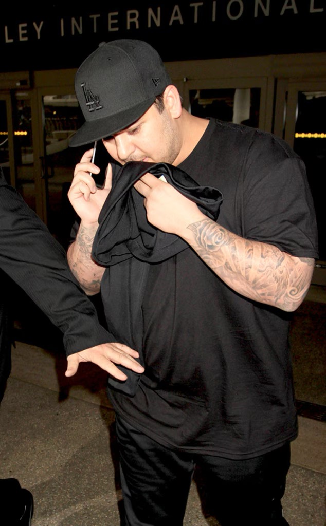 Rob Kardashian is photographed outside an airport, holding a black shirt over part of his face while talking on his phone. The image recalls his difficult relationship with paparazzi attention, which he later revealed had contributed to his decision to miss Kim Kardashian's 2014 wedding. | Source: Getty Images.