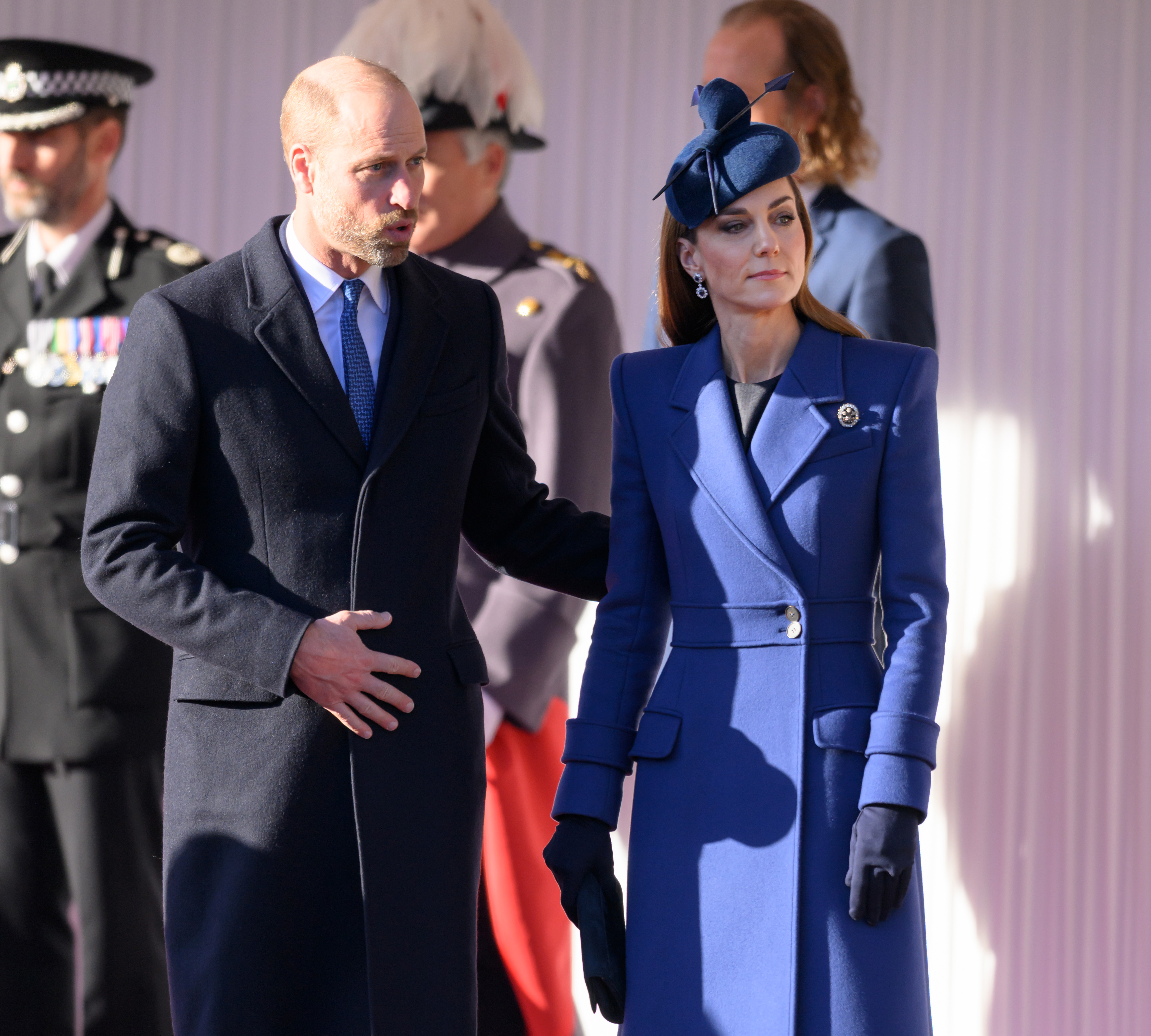Prince William and Catherine, Princess of Wales, stand together during the ceremonial welcome at Windsor Castle on December 3, 2025, marking the start of the German State Visit. Catherine looks tailored and polished in a striking cobalt-blue coatdress with structured shoulders, notch lapels, and a belted waist, accessorized with a matching sculptural bow fascinator, dark blue gloves, and a historic diamond brooch, standing beside William, who is dressed in a dark overcoat and blue tie. | Source: Getty Images