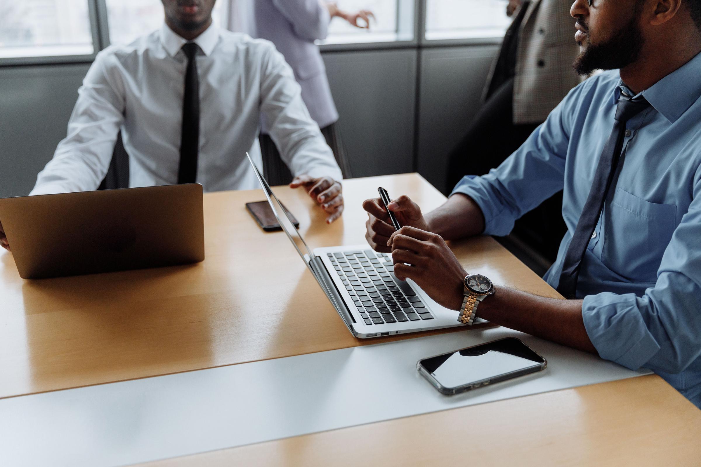 Two men in dress shirts and ties sit at a conference table with laptops and phones. | Source: Pexels