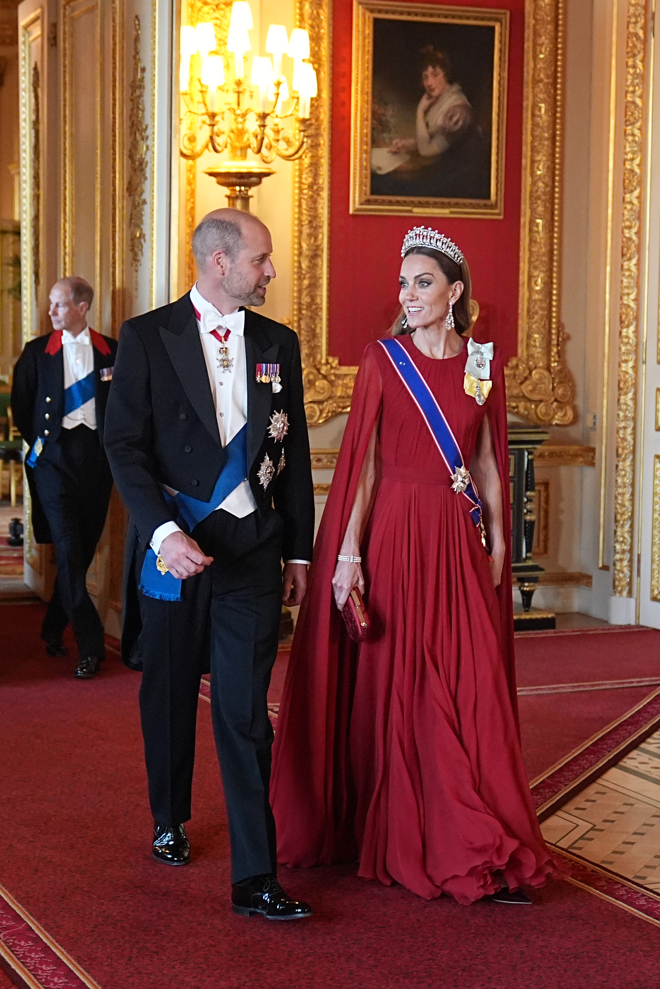 Prince William and Catherine, Princess of Wales, make a grand entrance for the state banquet honoring French President Emmanuel Macron and First Lady Brigitte Macron at Windsor Castle on July 8, 2025. Catherine commands attention in a pleated crimson-red cape gown paired with Queen Mary's Lover's Knot Tiara, diamond drop earrings, the Royal Victorian Order sash, and King Charles III's Royal Family Order, smiling alongside William, who wears a formal Windsor tailcoat with white tie and full ceremonial honors. | Source: Getty Images