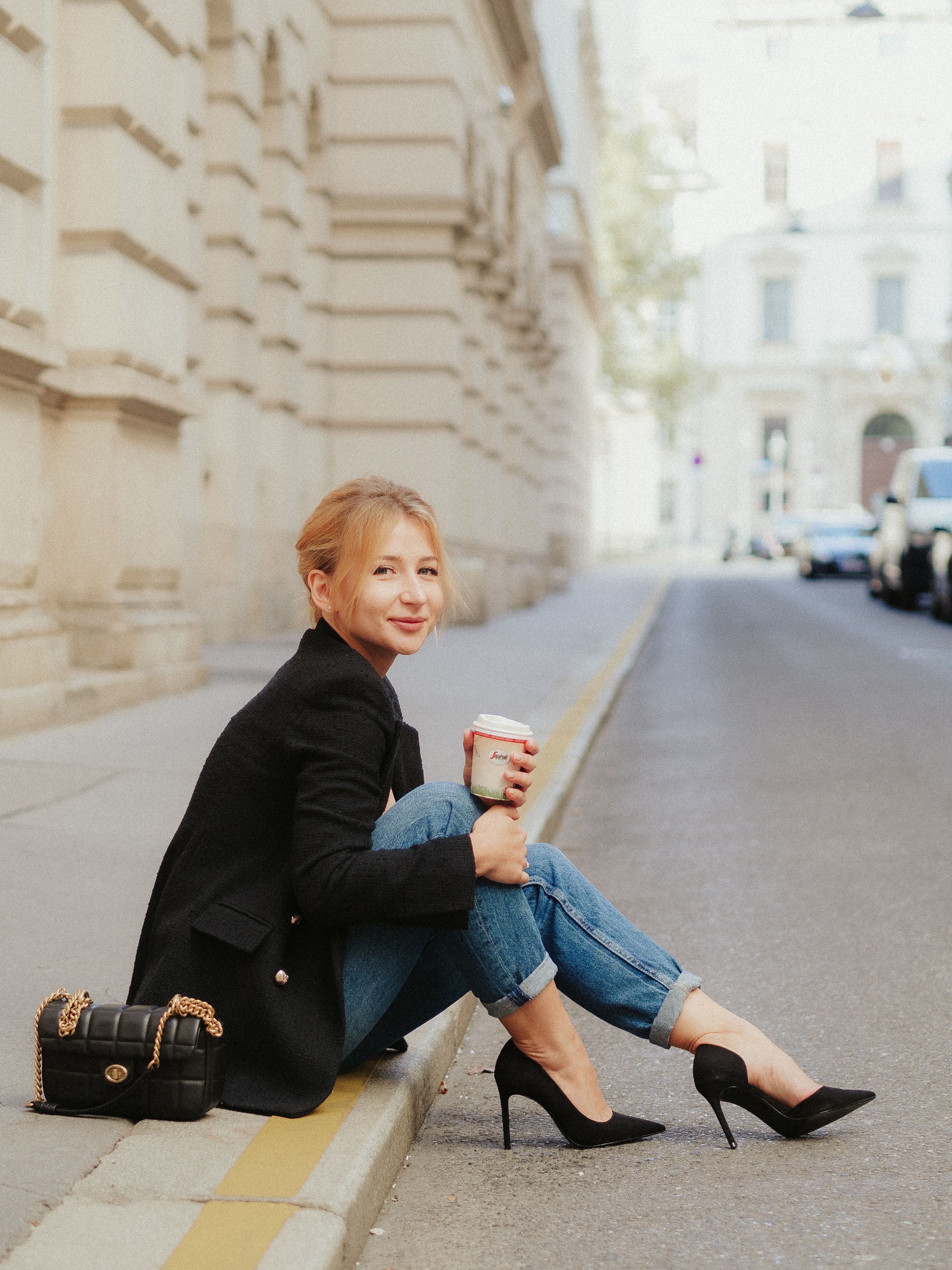 A woman seated on a sidewalk dressed in a black blazer, jeans, and a pair of black high-heeled shoes | Source: Pexels
