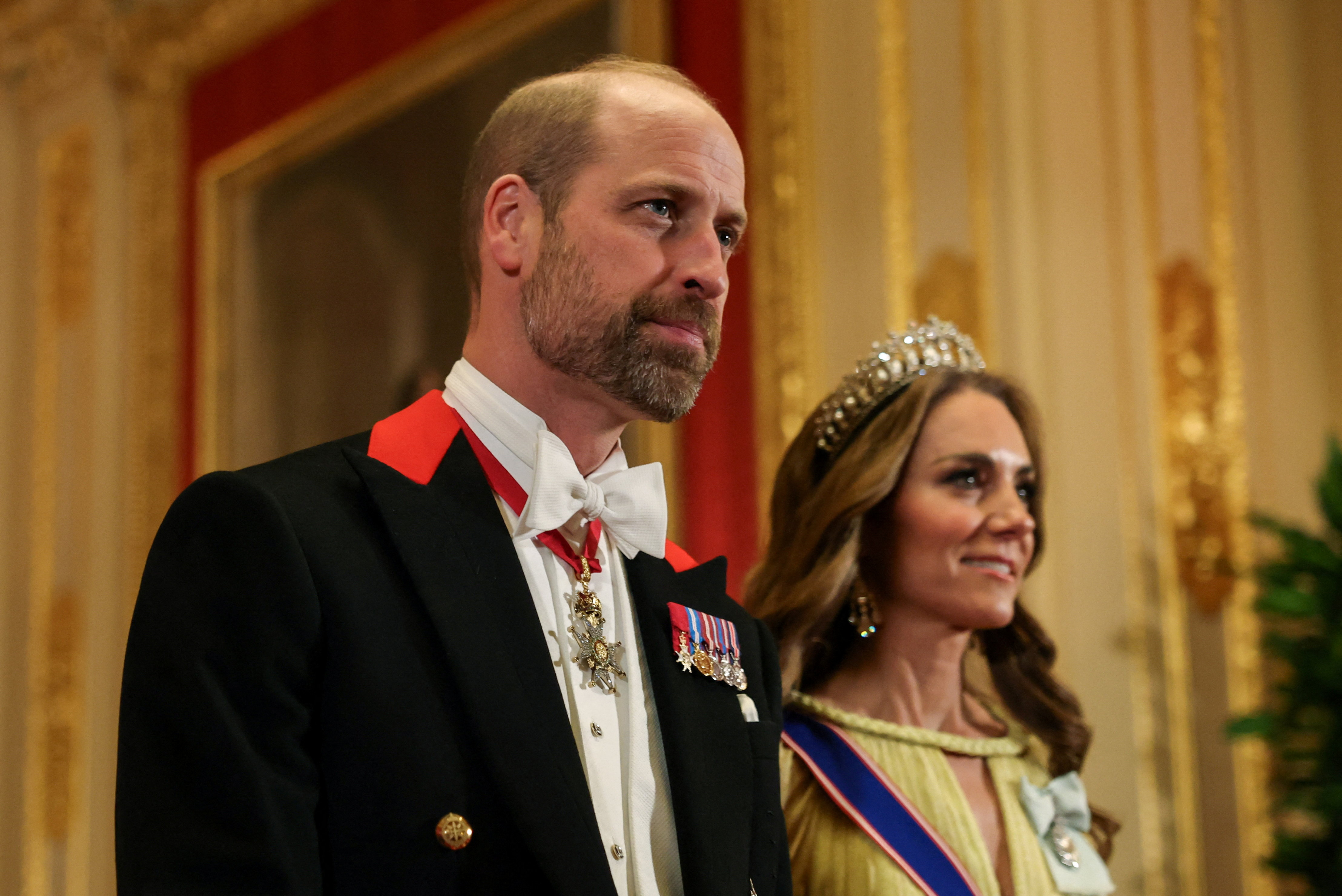 Prince William shows off noticeable facial scruff alongside Catherine, Princess of Wales, during their evening entrance at Windsor Castle on October 20, 2026. The future king pairs his trimmed beard with a traditional black Windsor tailcoat featuring vibrant red lapel accents, white tie, and miniature medals, while Catherine looks on in her tiara and pastel-yellow gown. | Source: Getty Images