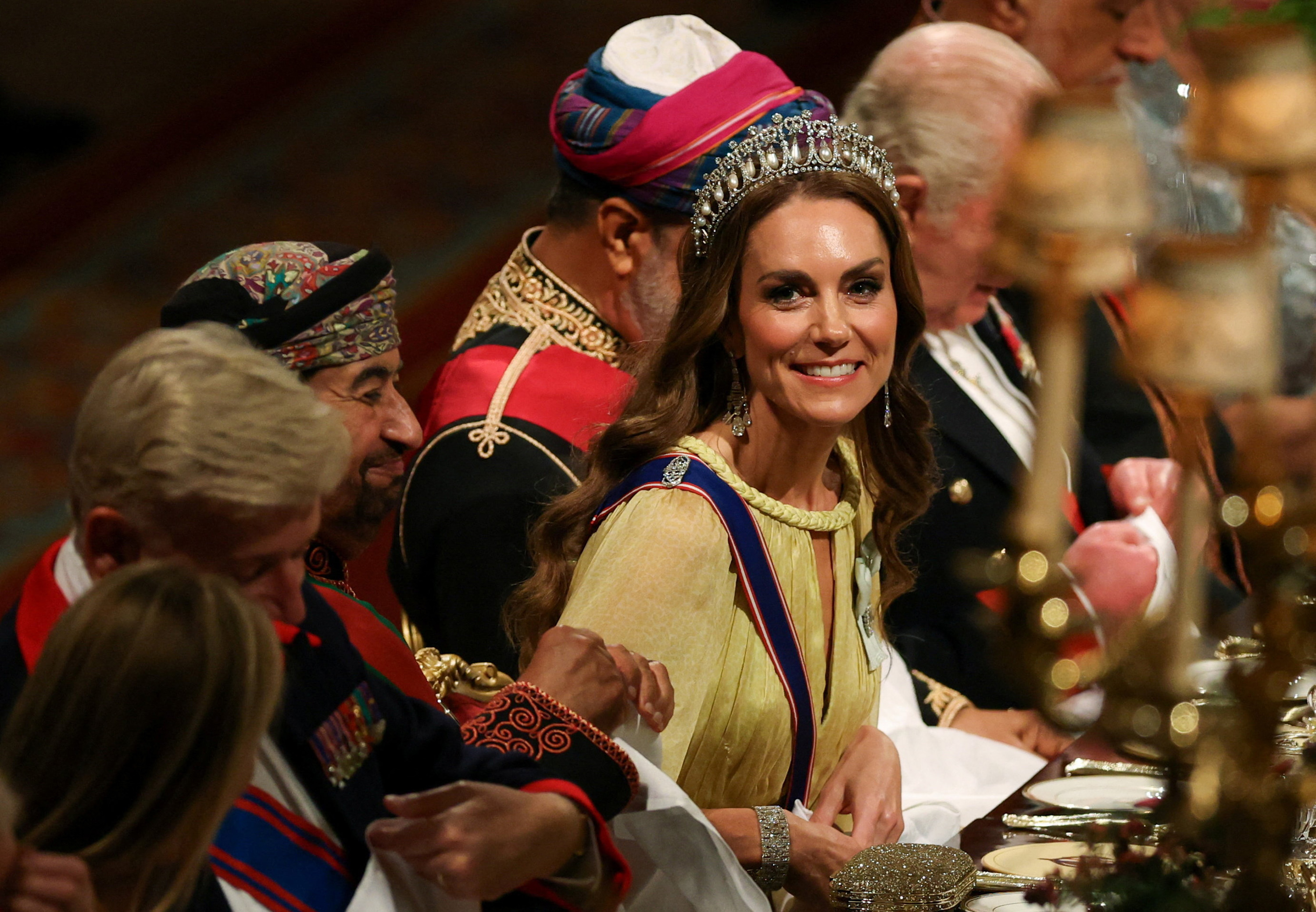 Seated along the banquet table in St. George's Hall on October 20, 2026, Catherine, Princess of Wales, beams warmly as dinner proceedings begin. Her radiant beauty look features soft brunette waves cascading over her shoulders beneath the pearl-and-diamond Lover's Knot Tiara, paired with diamond chandelier earrings, smokey eye makeup, and the braided rope neckline of her yellow cape gown. | Source: Getty Images
