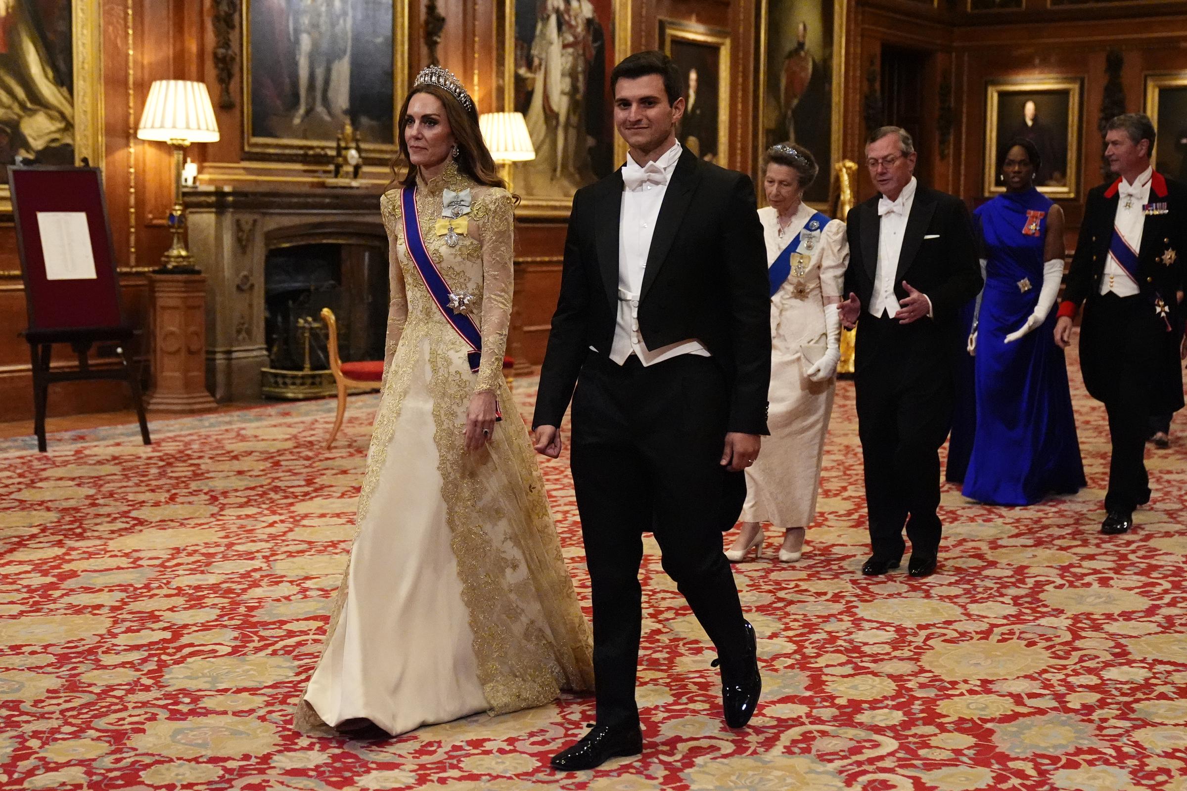 Catherine, Princess of Wales, walks alongside Michael Boulos during the formal procession into the state banquet at Windsor Castle on September 17, 2025, held in honor of U.S. President Donald Trump and First Lady Melania Trump. Catherine stuns in an ivory-and-gold lace gown featuring long illusion sleeves and intricate gold embroidery, accessorized with Queen Mary's Lover's Knot Tiara, the Royal Victorian Order sash, and King Charles III's Royal Family Order, while Boulos is sharply dressed in a black tailcoat and white tie. | Source: Getty Images