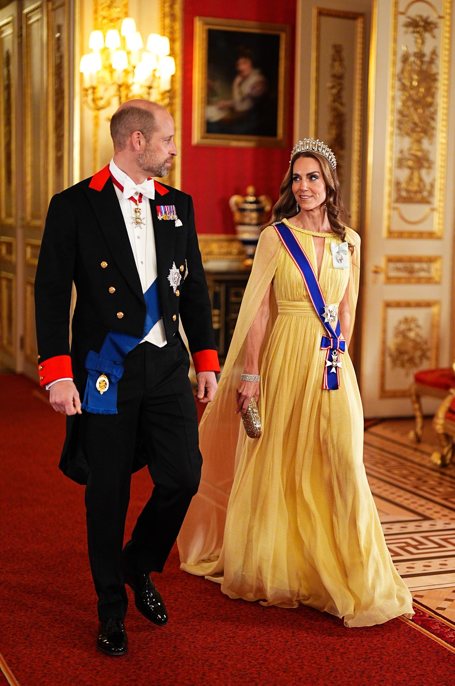 Walking side-by-side along the red carpeted corridors of Windsor Castle on October 20, 2026, the Prince and Princess of Wales display regal elegance ahead of the evening's formal dinner. Catherine glides in a pleated pastel-yellow cape gown by Elie Saab, carrying a sparkling gold evening clutch, while Prince William sports a well-fitted tailcoat, white waistcoat, and bow tie alongside his royal garter decorations. | Source: Getty Images