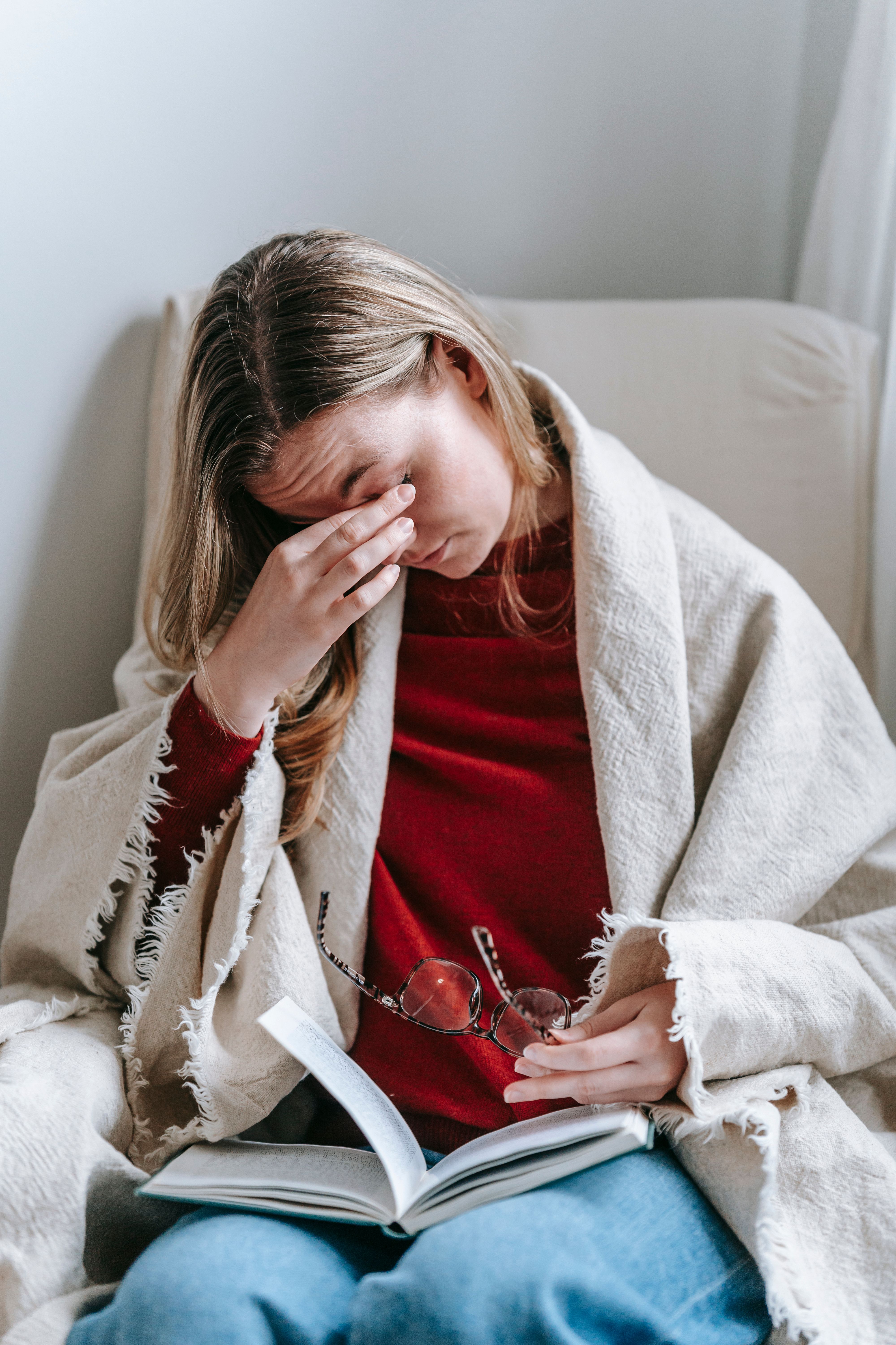 A woman rubbing her eyes while seated on a hair with a book on her lap | Source: Pexels