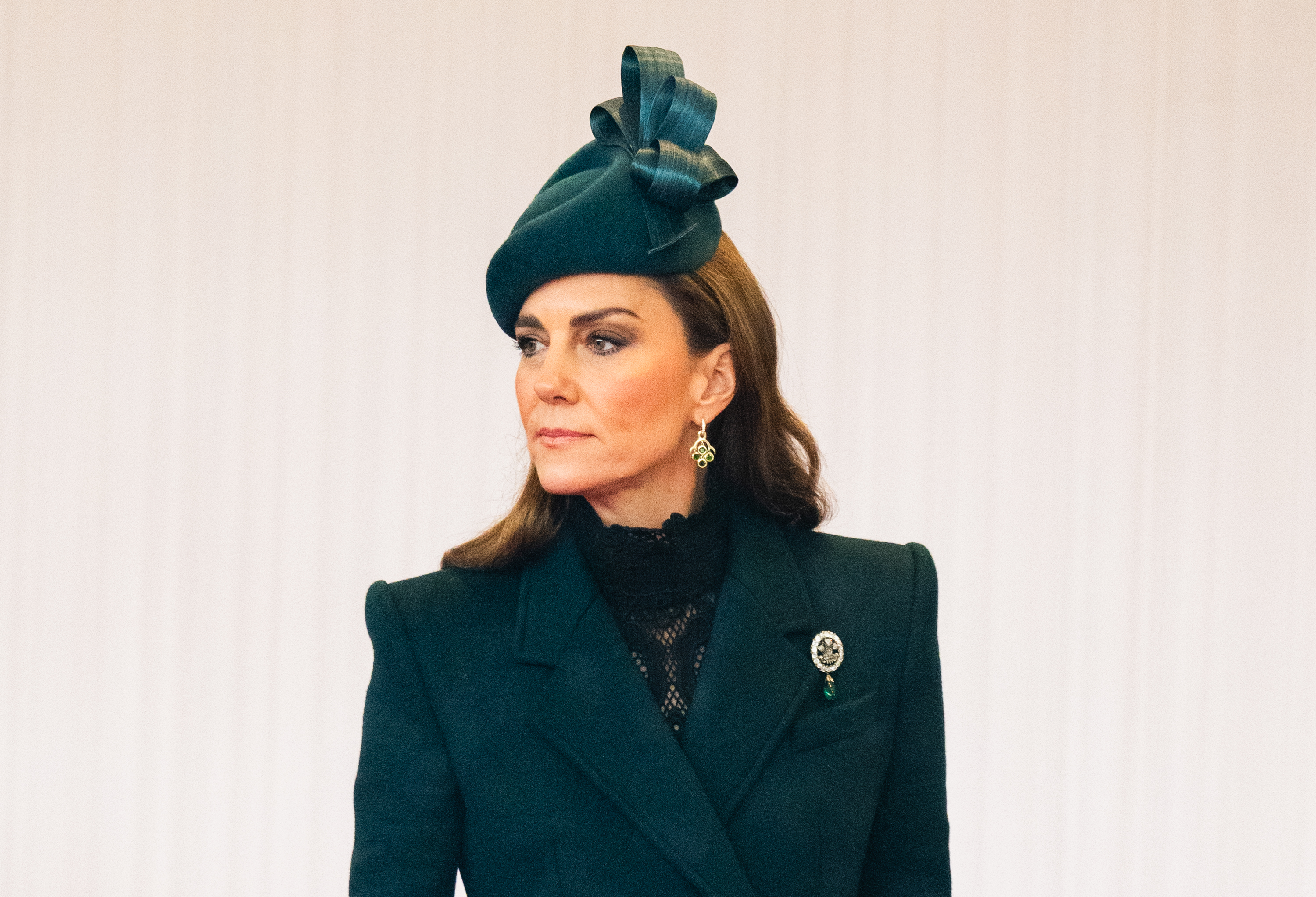 A close-up portrait shows Catherine, Princess of Wales, on October 20, 2026, showcasing the intricate styling of her daytime look. Her tailored notch-lapel coat frames a sheer black lace high collar, accompanied by a sculptural green bow fascinator, Cassandra Goad chandelier earrings, and the sparkling Prince of Wales Feathers Brooch pinned to her left shoulder. | Source: Getty Images