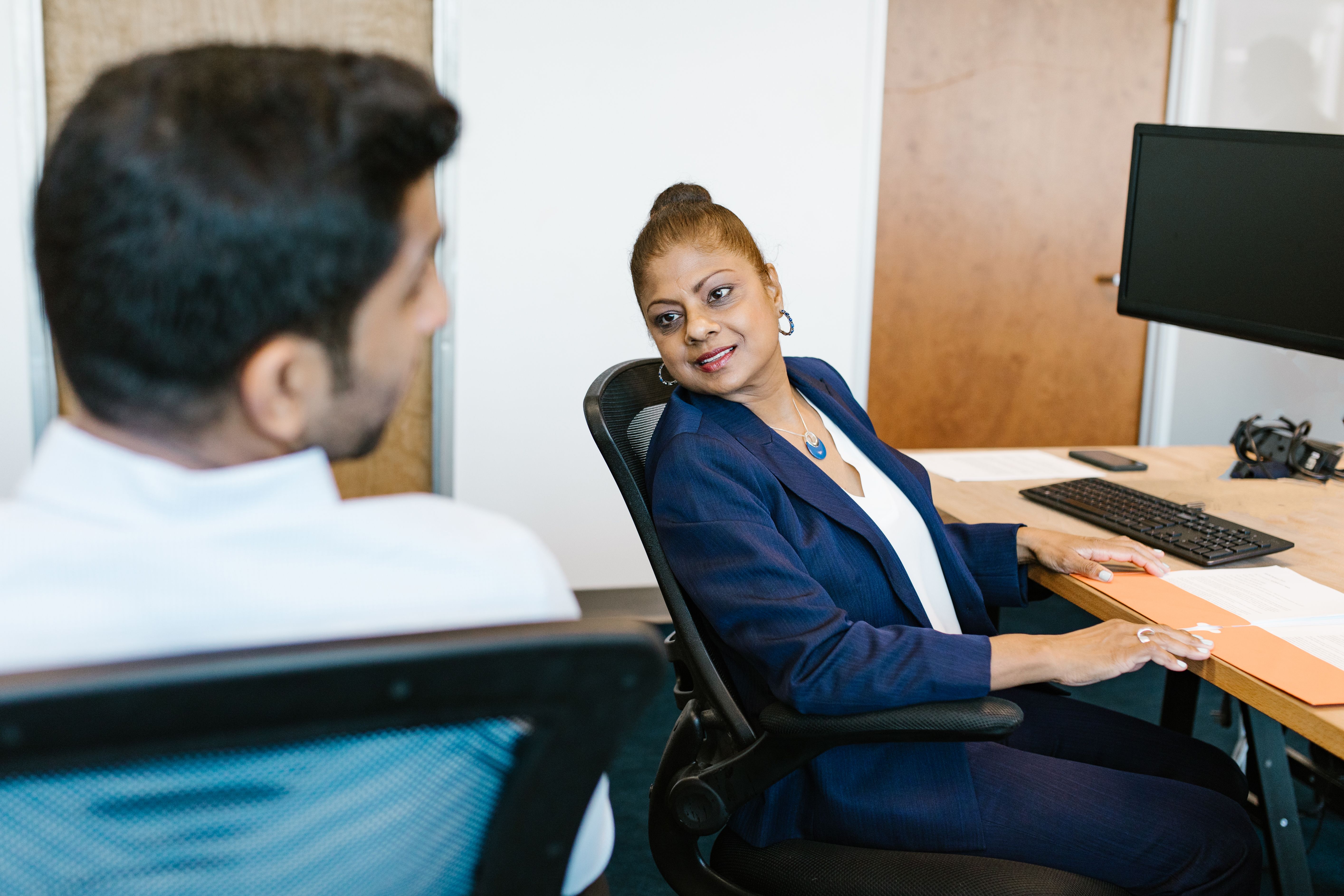A woman in a navy blazer sits at a desk and looks toward a man seen from behind. | Source: Pexels