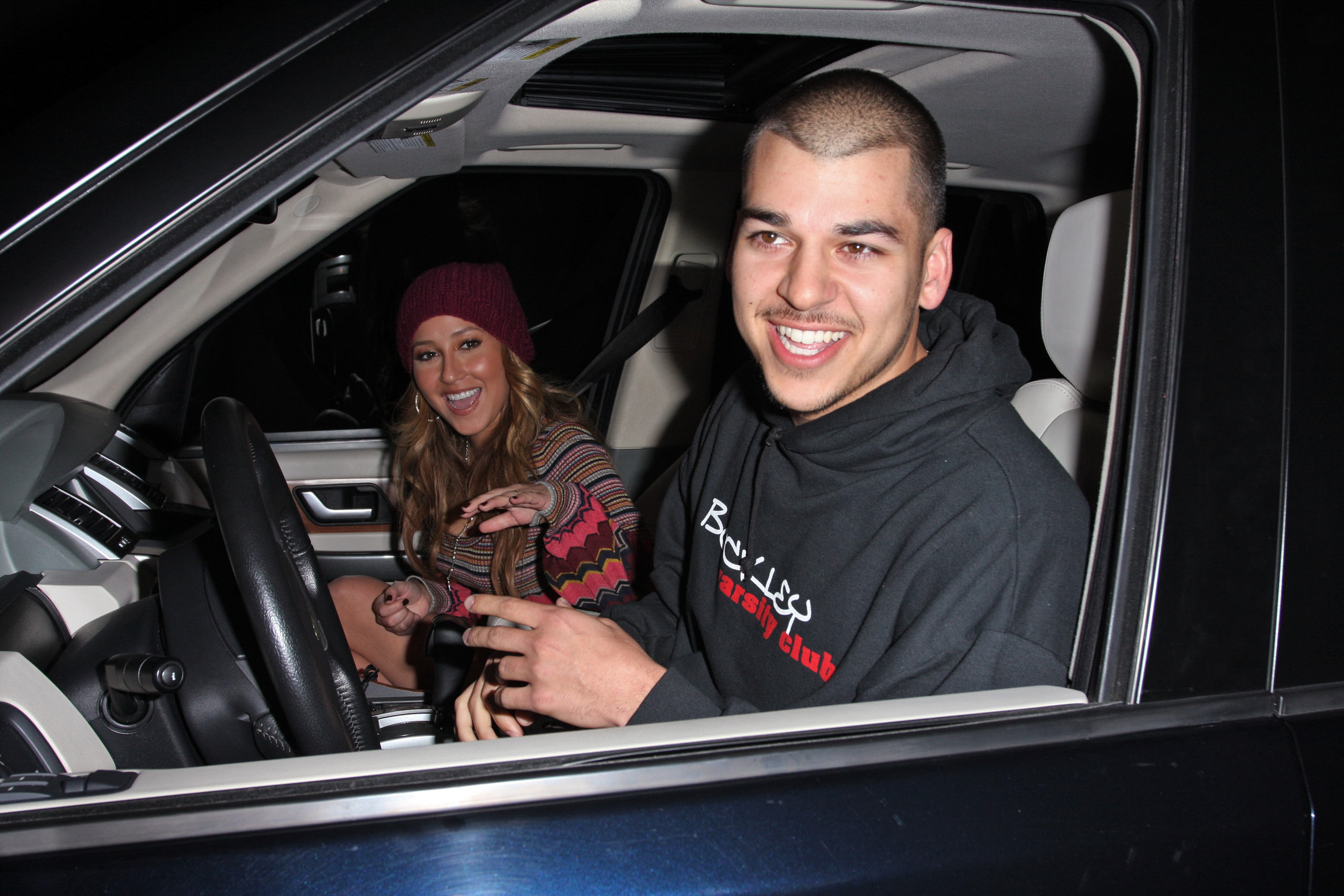Rob Kardashian smiles behind the wheel while a woman sitting beside him waves toward photographers during a nighttime outing. The photo captures a relaxed, cheerful moment from his earlier years in the spotlight. | Source: Getty Images.