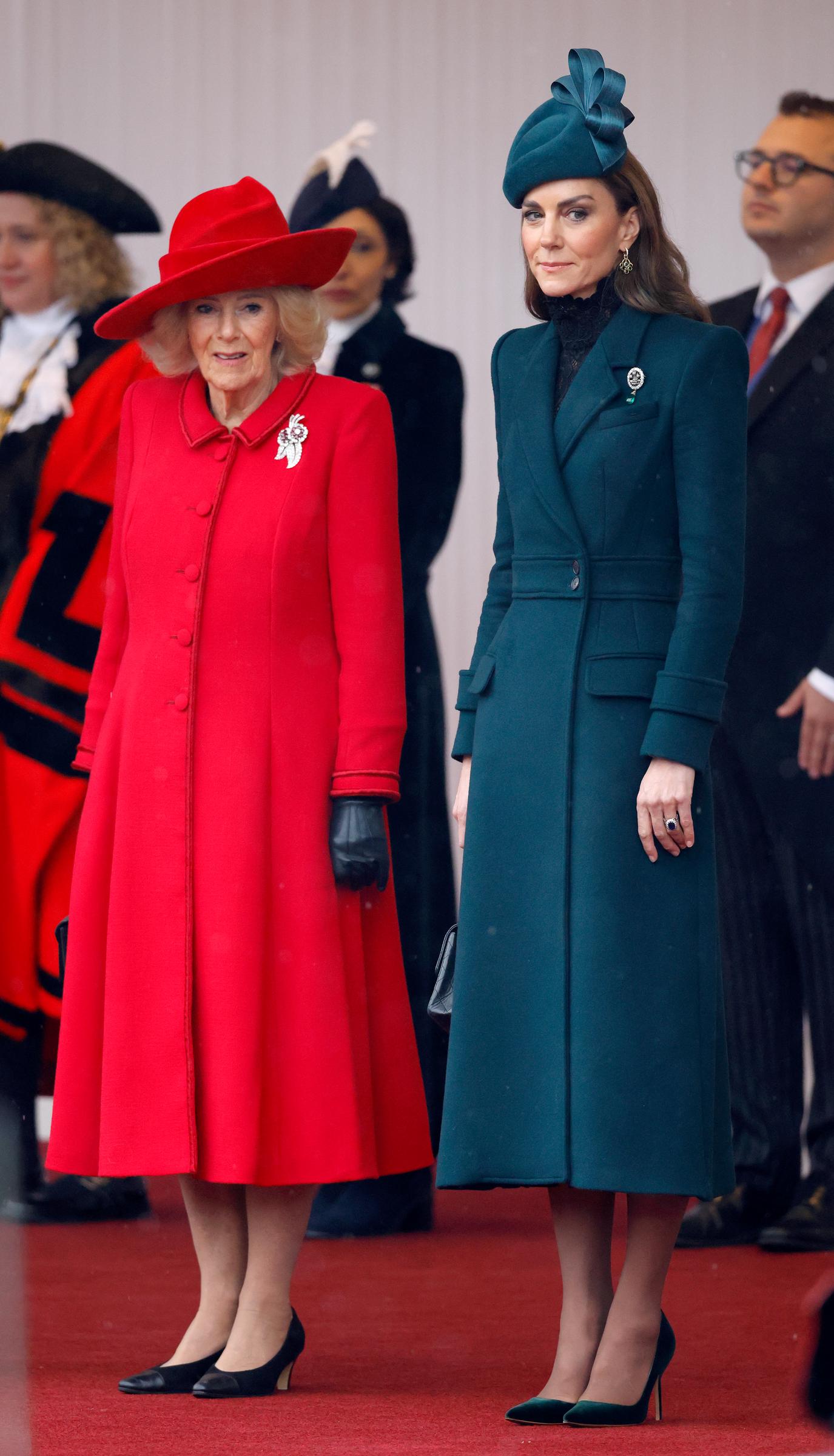 Catherine, Princess of Wales, stands on the red carpet alongside Queen Camilla in Windsor on October 20, 2026. Offering a striking contrast to Camilla’s vibrant red flared coat and matching brimmed hat, Catherine wears an emerald-green midi coatdress featuring sharp structured shoulders, paired with a matching sculpted fascinator, black lace detailing at the neckline, and green velvet pointed-toe heels. | Source: Getty Images