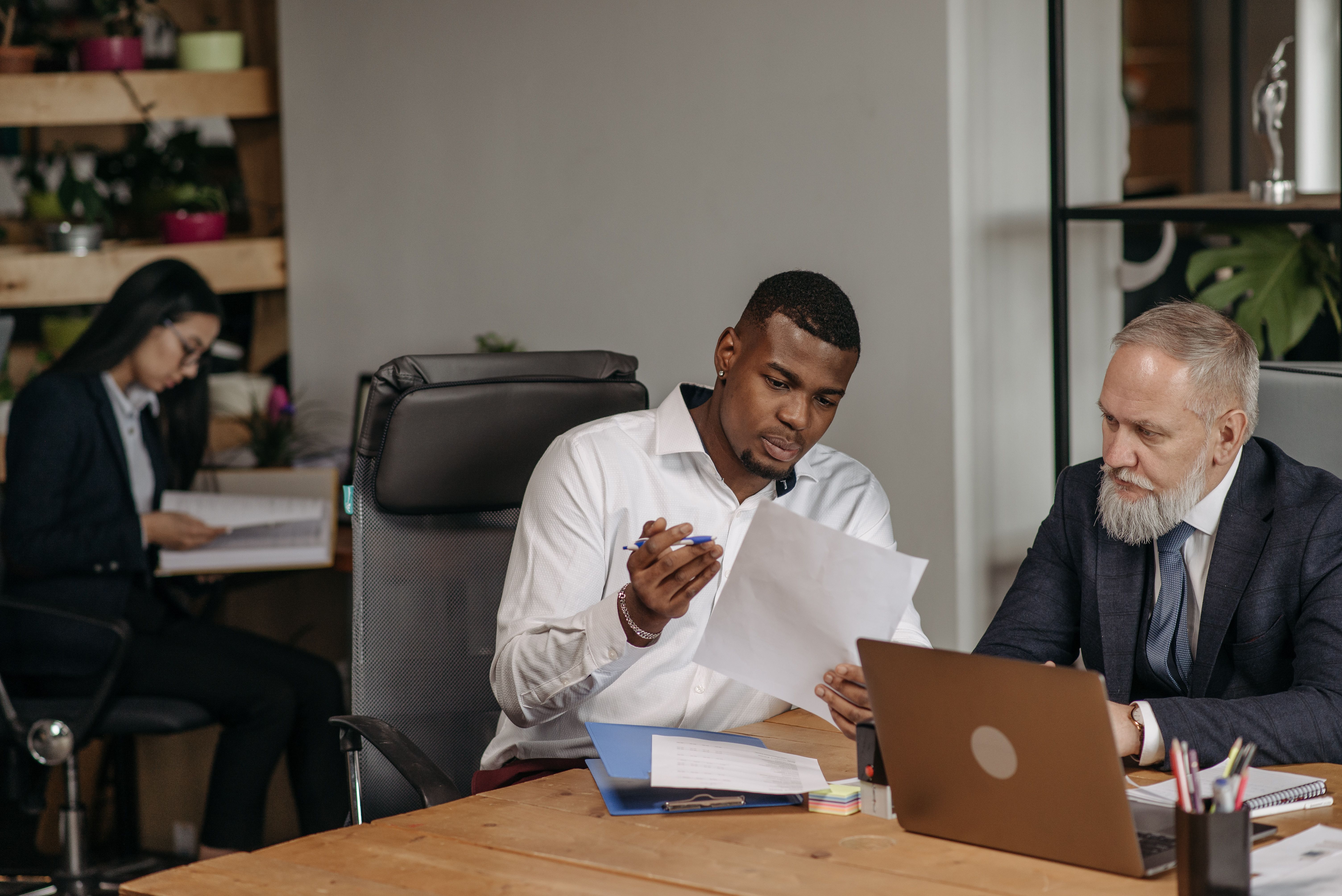 Two men sit at a desk and look at a sheet of paper, with a laptop and folders in front of them. | Source: Pexels
