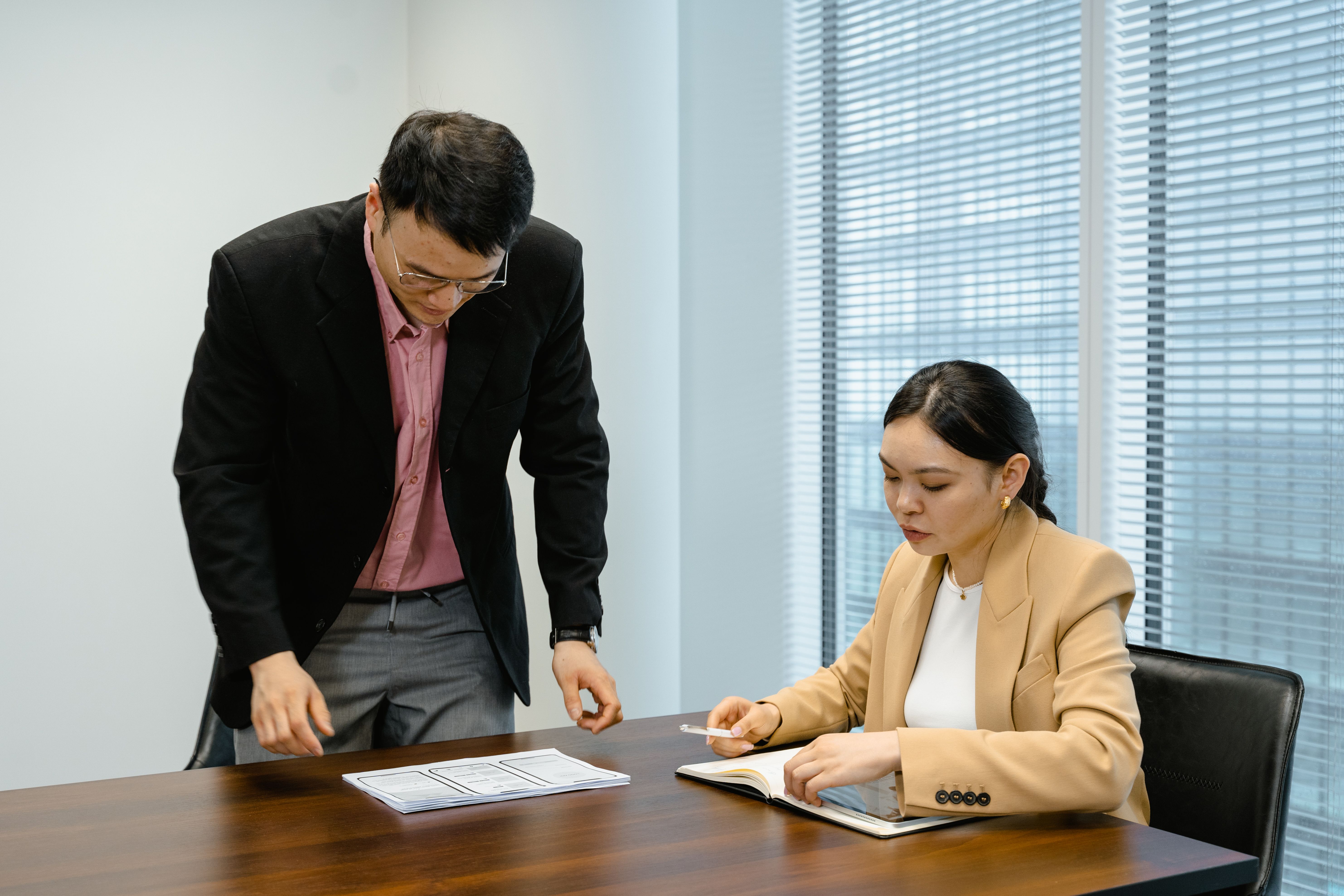 A man leans over a desk of papers while a woman in a tan blazer writes in a notebook. | Source: Pexels