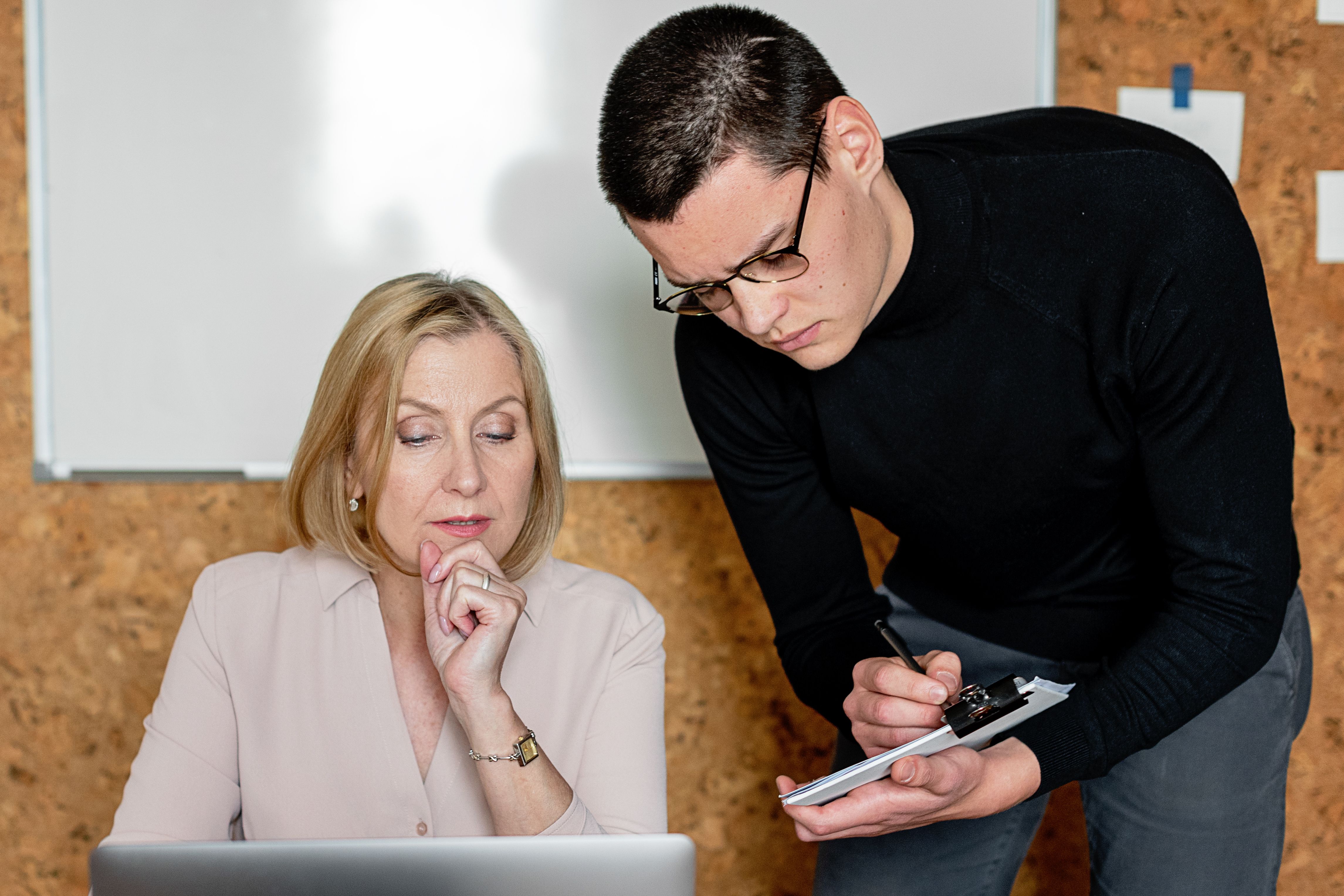 A man leans over a seated woman and writes on a clipboard while she looks at a laptop. | Source: Pexels