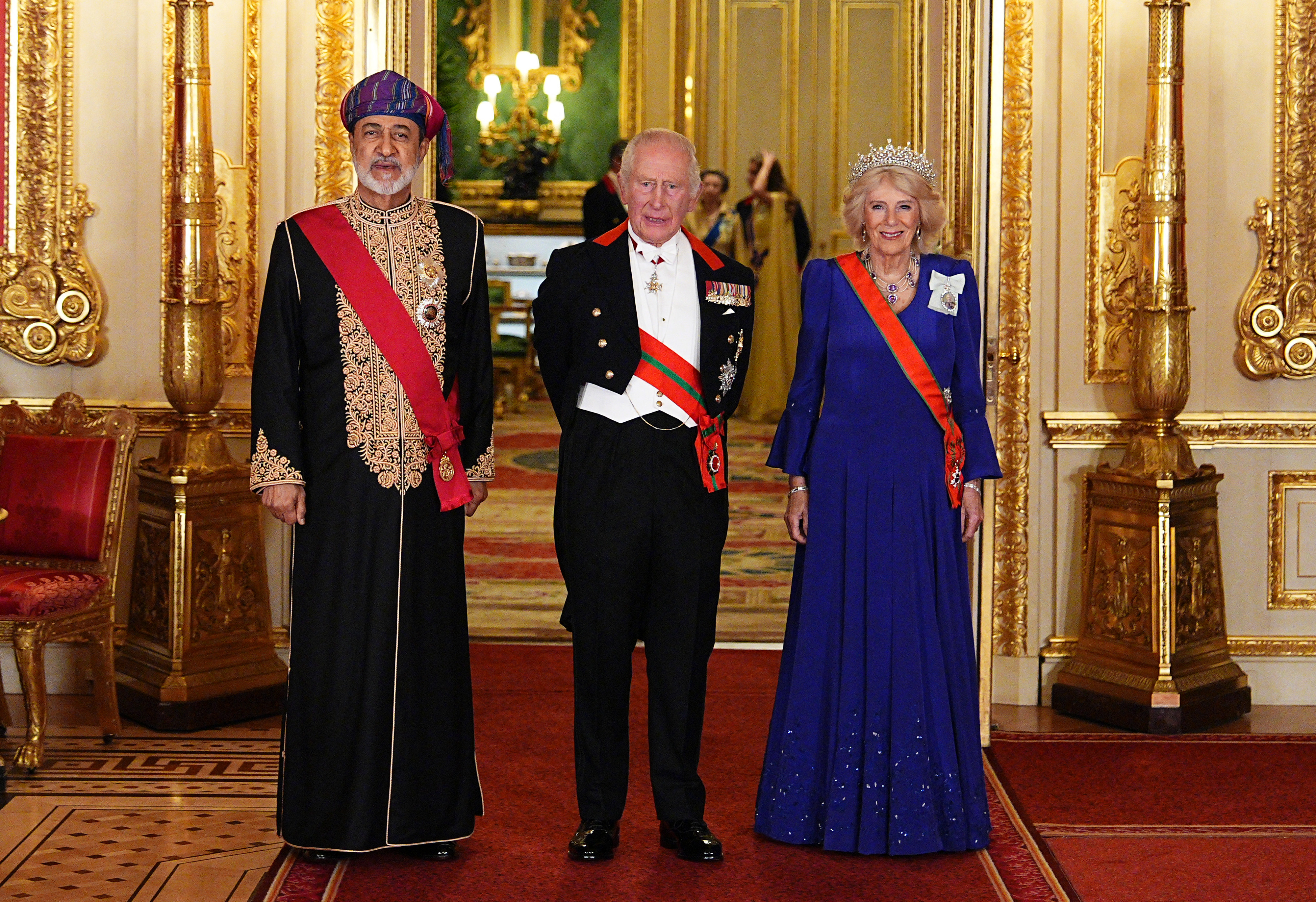 Sultan Haitham bin Tarik of Oman poses alongside King Charles III and Queen Camilla inside Windsor Castle ahead of the lavish State Banquet on October 20, 2026, in Windsor, England. Flanked by ornate gilded hall decor, King Charles III wears a formal evening tailcoat with military medals and order sashes alongside the Sultan, who dons an intricately gold-embroidered black formal robe, red ceremonial sash, and colorful traditional massar turban, while Queen Camilla shines in a blue pleated gown crowned by the sparkling Belgian Sapphire Tiara and an orange order sash. | Source: Getty Images
